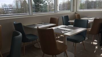 A dining area with tables and chairs, large windows with a view of trees and buildings, and a wooden floor.