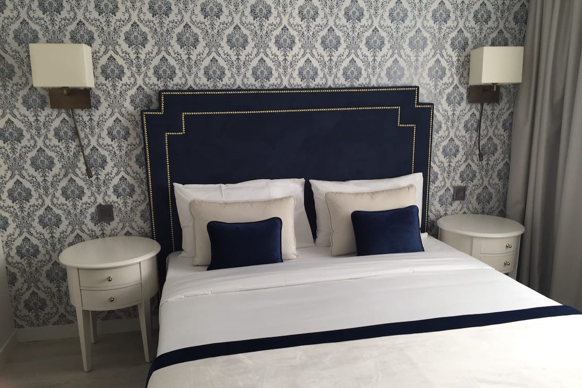 Een netjes opgemaakt bed met een marineblauwe hoofdbord, witte beddengoed en twee bijpassende marineblauwe kussens. Er staan twee nachtkastjes met lampen, en de muren hebben behang met een patroon.