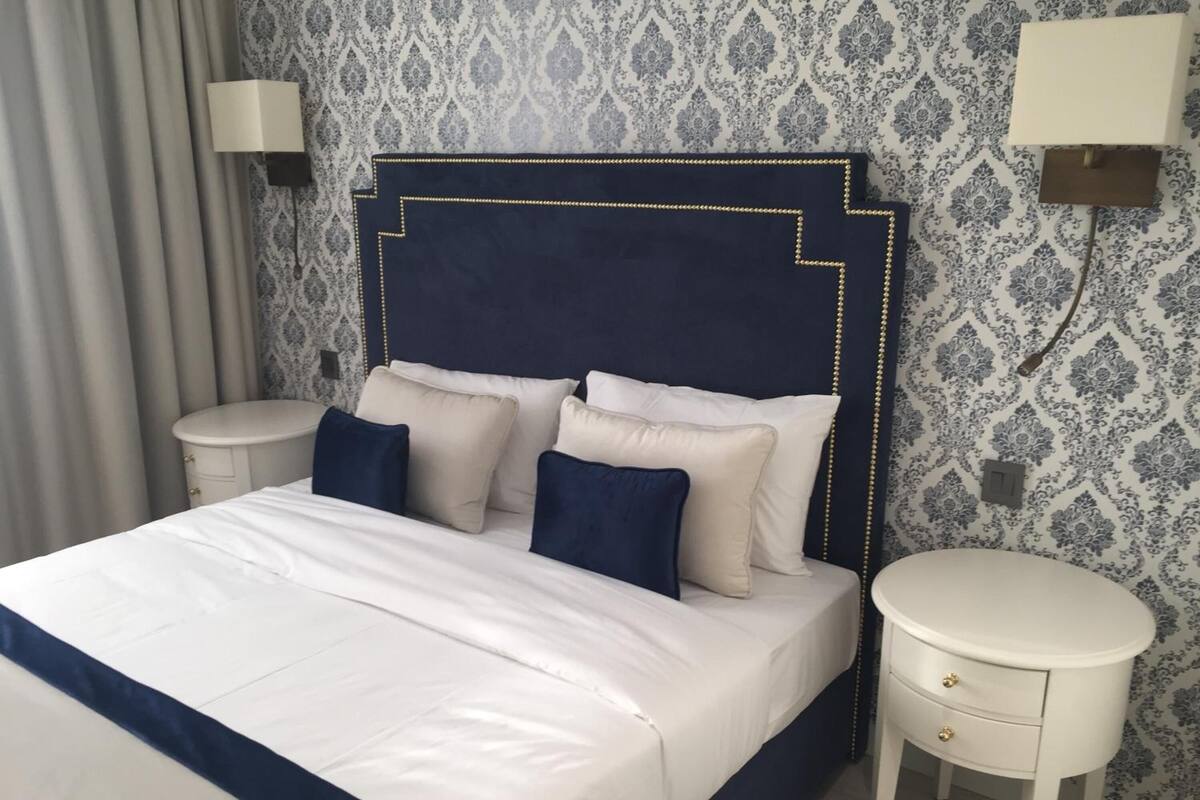 Een netjes opgemaakt bed met wit en marineblauwe beddengoed, een donker hoofdbord met gouden details en twee nachtkastjes met lampen.