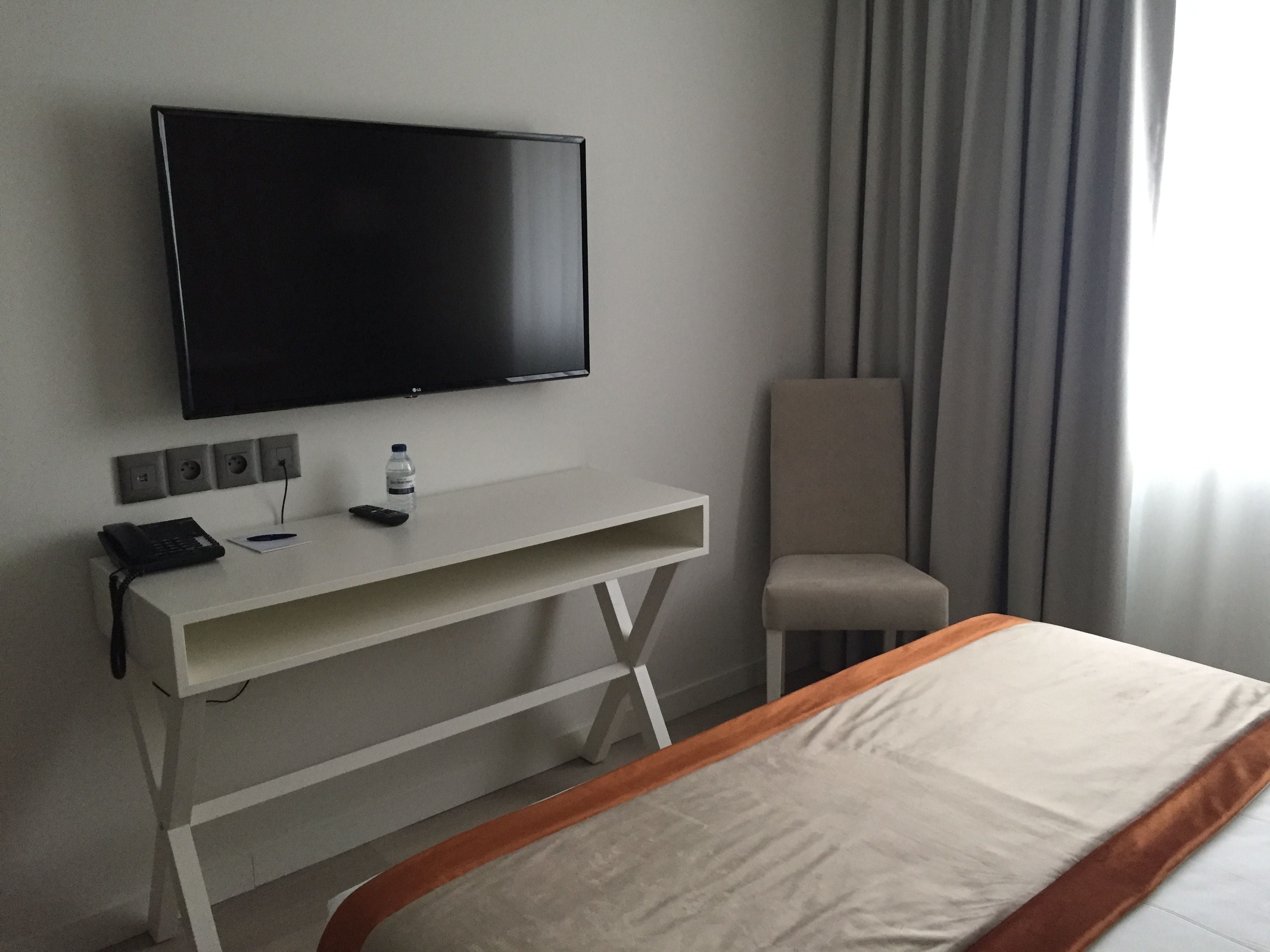 A hotel room with a bed, a TV mounted on the wall, a desk with a phone and remote control, a chair, and a window with curtains.