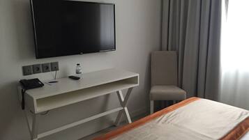 Een hotelkamer met een bed, een aan de muur bevestigde televisie, een bureau met telefoon en afstandsbediening, een stoel en een raam met gordijnen.