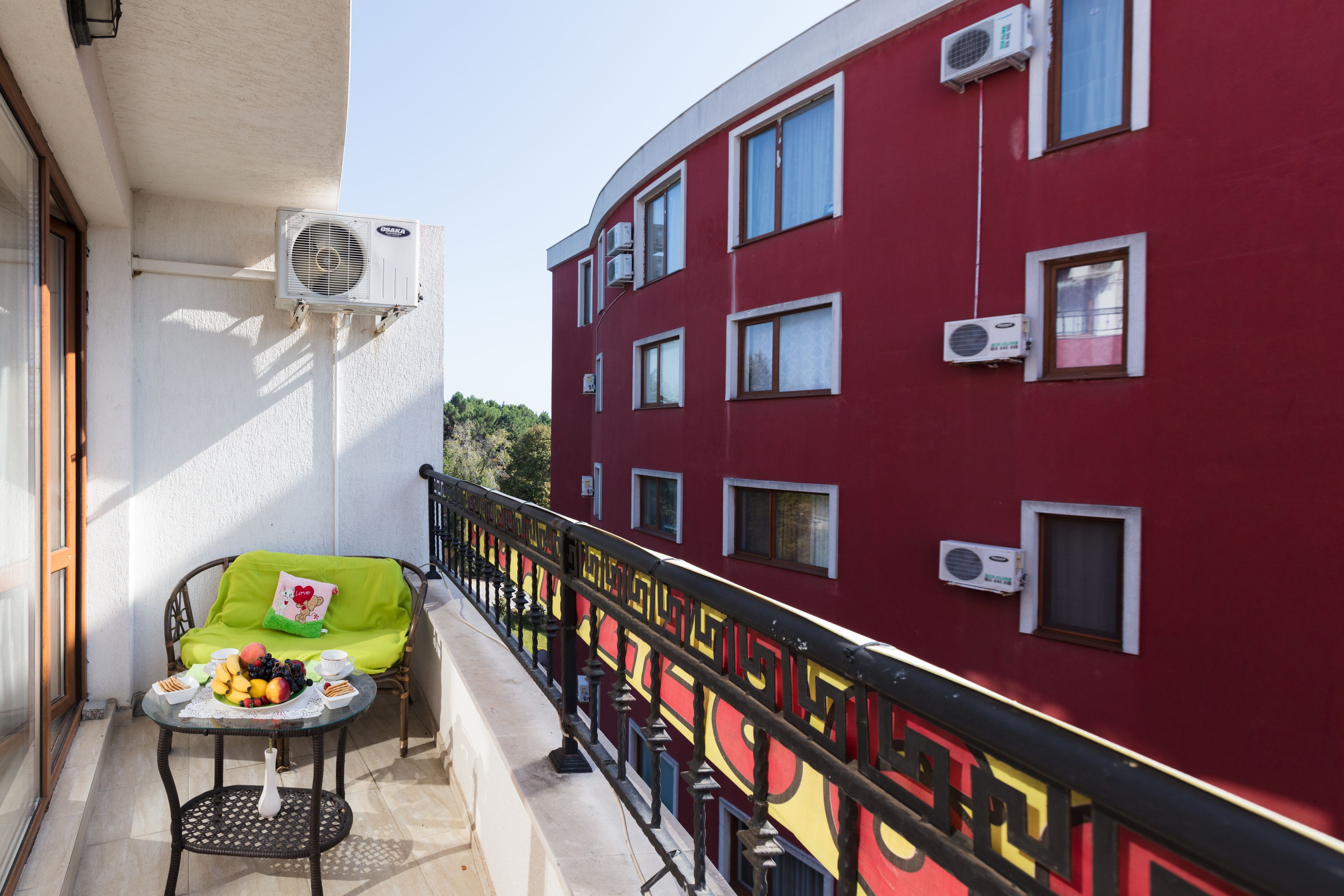 Ein Balkon mit Frühstückstisch, einem grünen Stuhl und einem roten Gebäude mit vielen Fenstern und Klimaanlagen.