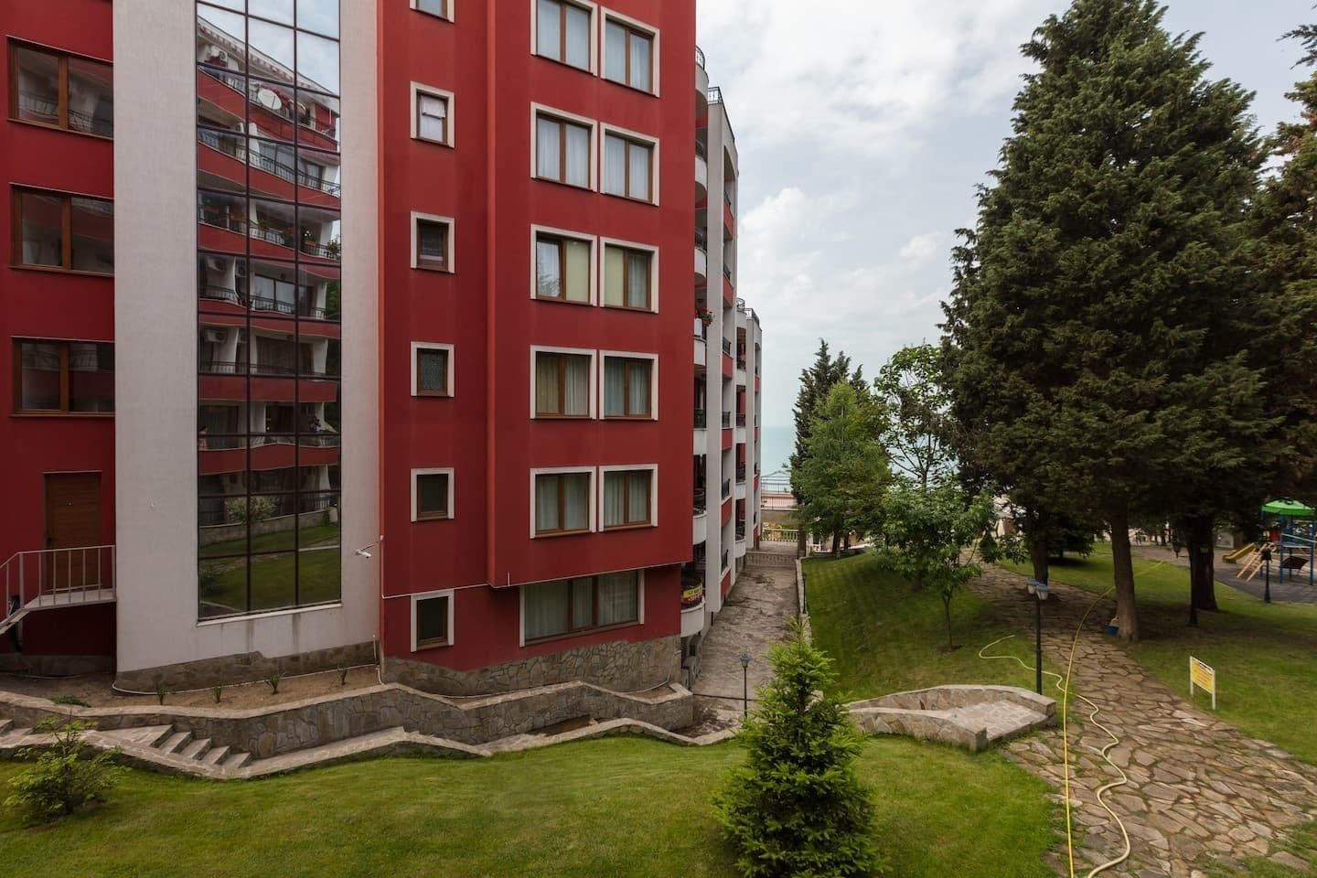 Een rood meerlaags gebouw met balkons, een pad van stenen en een park met bomen en een speeltuin.