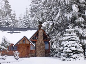 Une cabane en rondins avec une cheminée en pierre, entourée d’arbres couverts de neige.