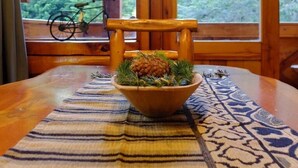 Une table à manger en bois, surmontée d’un set de table en fibres tressées et agrémentée d’un bol rempli de pommes de pin et de verdure. On aperçoit un vélo à travers une fenêtre au fond.