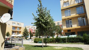 A courtyard with a tree, outdoor seating, and multi-story buildings with balconies. - Quiet Family Apartment in Sunny Day (Nessebar)