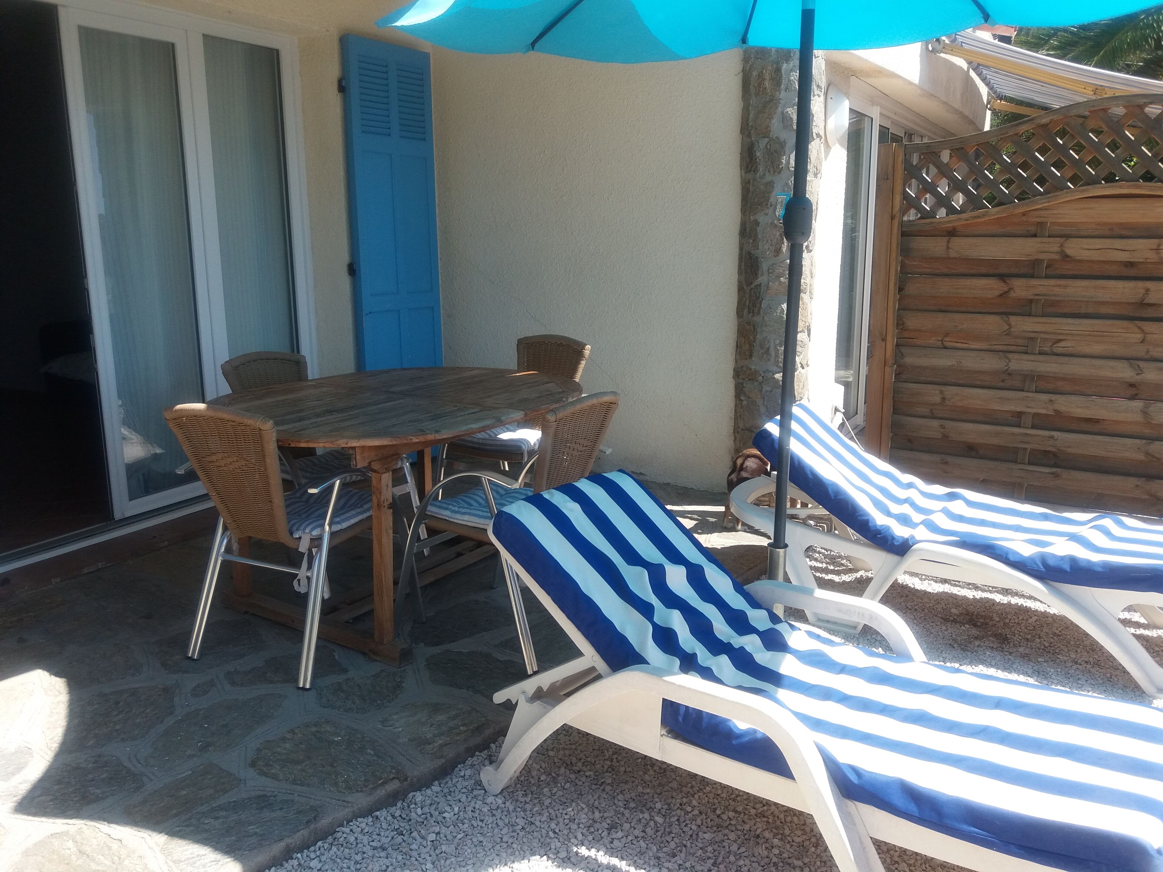 Eine Terrasse mit einem Holztisch, Stühlen und zwei Liegestühlen mit gestreiften Kissen sowie einem blauen Sonnenschirm.