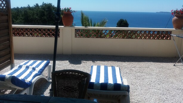 A balcony with two striped lounge chairs, a wicker chair, and a blue umbrella. - Exceptional sea view apartment in detached villa Côte d'Azur (Rayol-Canadel-Sur-Mer)