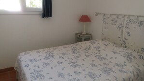 A bedroom with a bed, a nightstand with a lamp, a window with curtains, and a wall with a floral pattern. - Exceptional sea view apartment in detached villa Côte d'Azur (Rayol-Canadel-Sur-Mer)