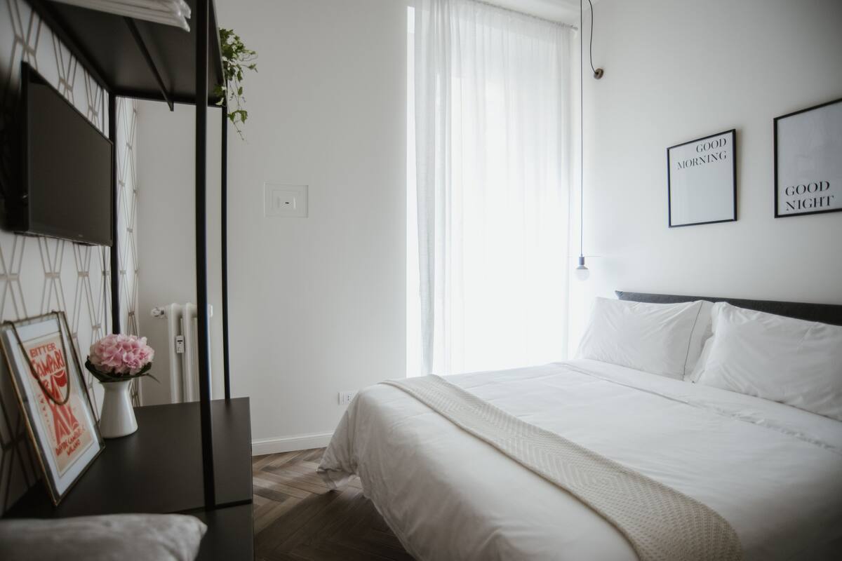A bedroom with a bed, a bedside table with a vase of flowers, a TV on the wall, and framed pictures.