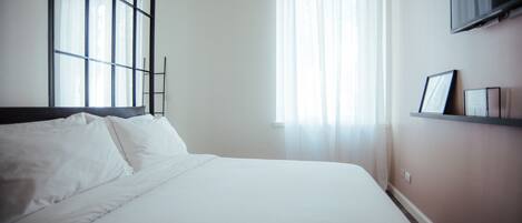 A neatly made bed in a room with a large window, a wall-mounted TV, and a shelf with a picture frame.
