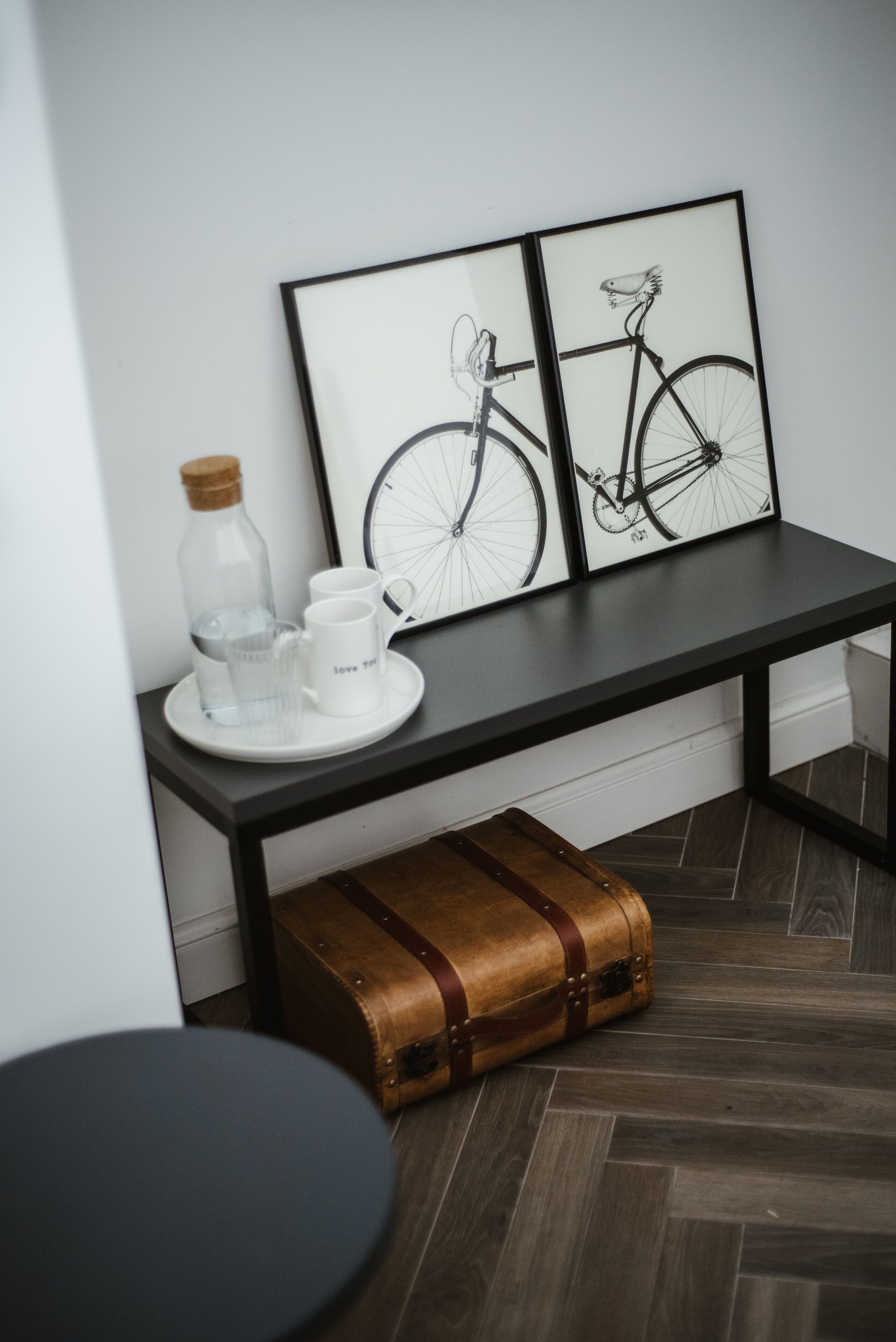 A room with a dark wooden floor, a black table with a white mug and glass, a cork bottle, and a brown suitcase.