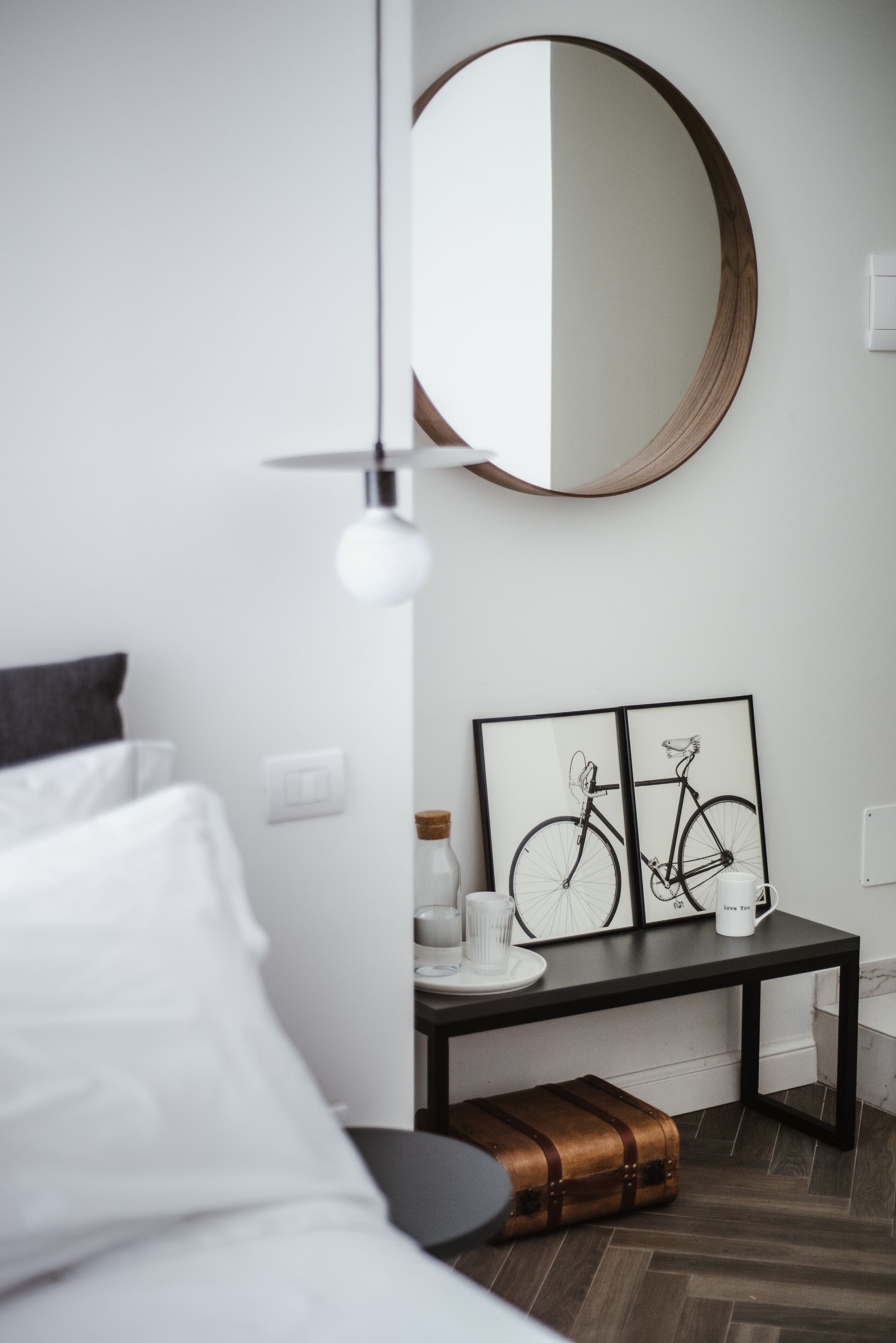 A modern bedroom with a bed, a bedside table with a framed bicycle artwork, a mirror, and a suitcase.