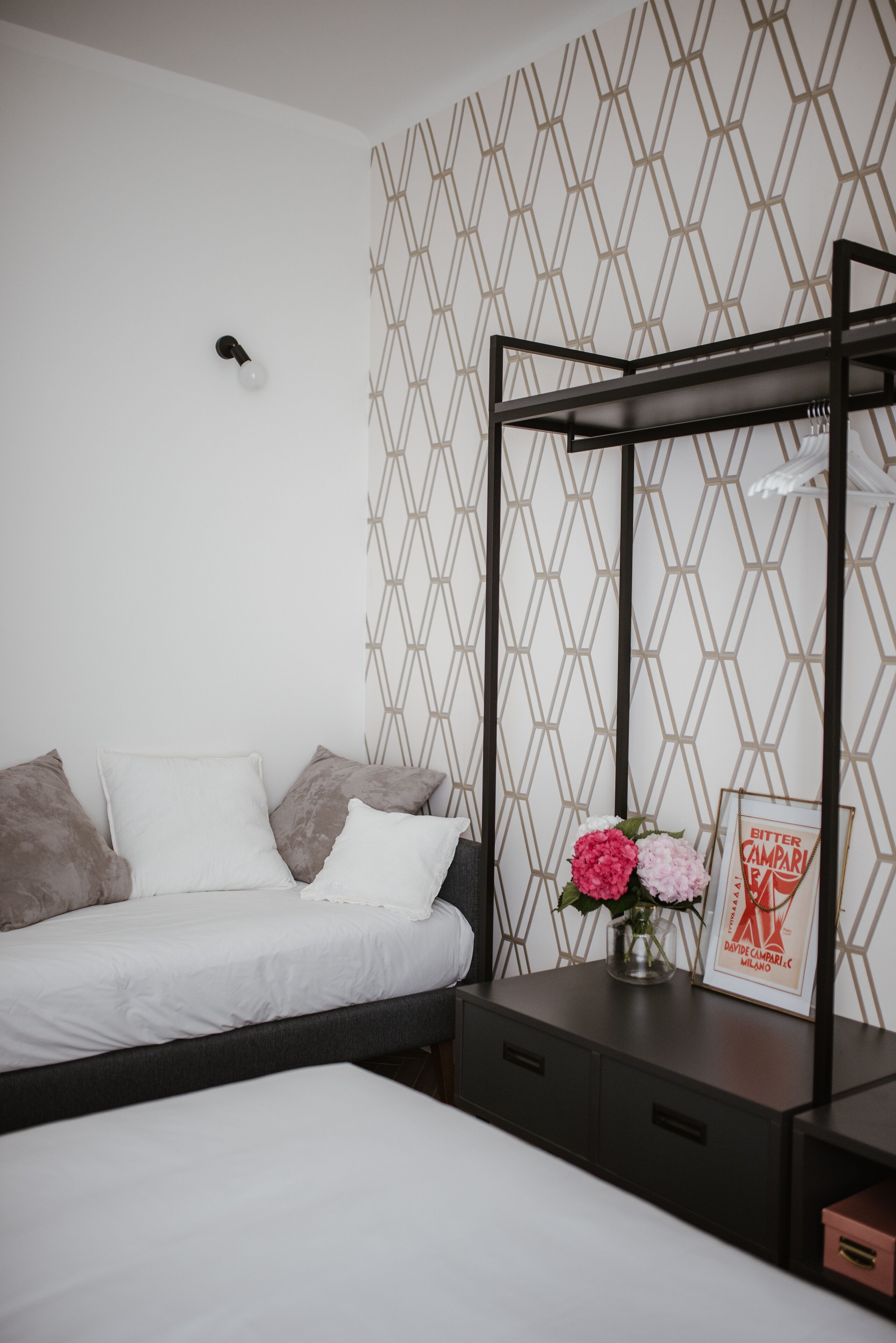A bedroom with a bed, a bedside table, a wall-mounted light, and a geometric-patterned wallpaper.