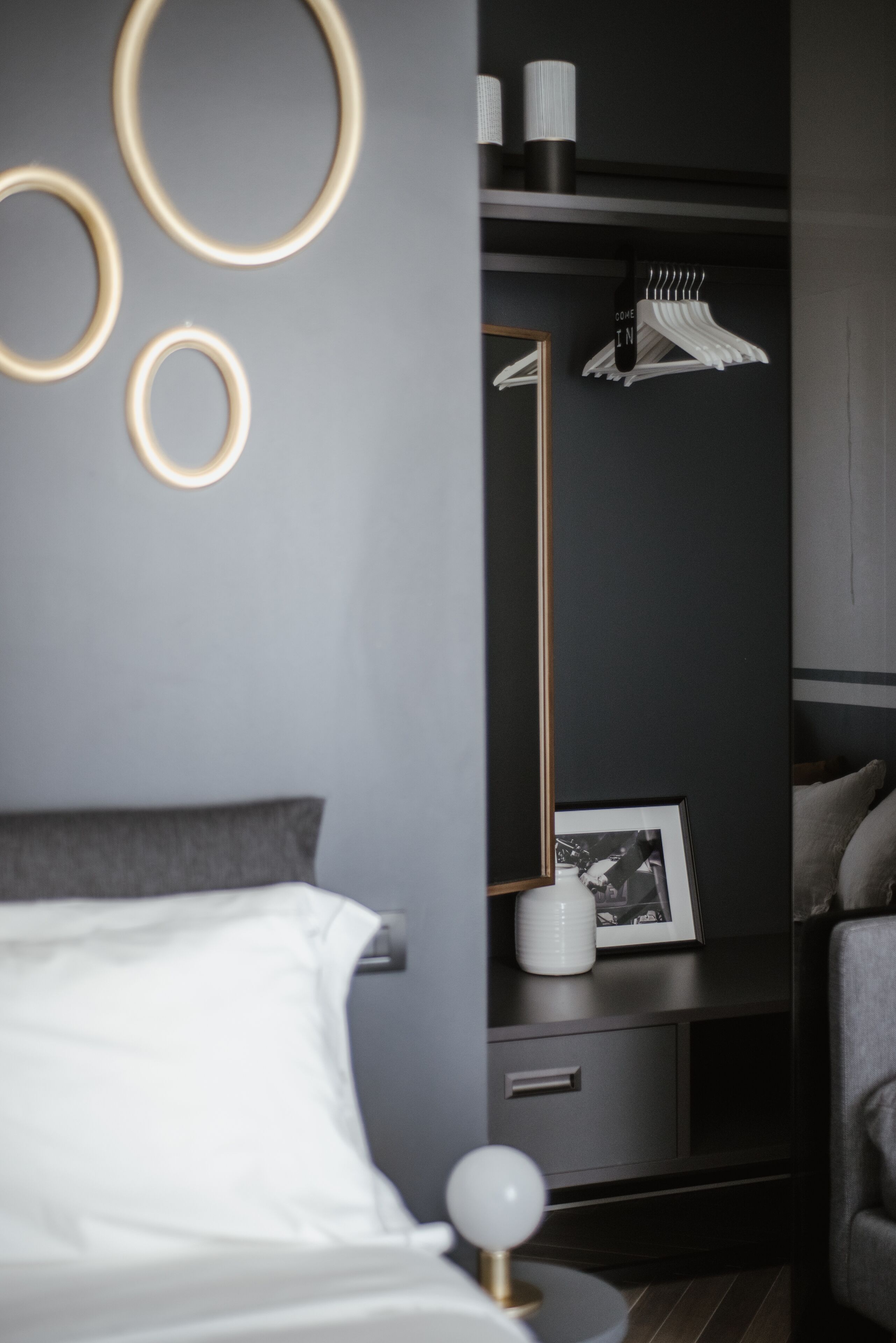 Een moderne slaapkamer met een bed, nachtkastje en een kledingkast met spiegel en hangende kleding.