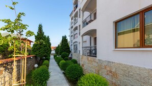A narrow pathway flanked by neatly trimmed hedges and a stone wall, leading to a multi-story building with balconies and a large window. - Apartment in Vista Del Mar (Ravda)