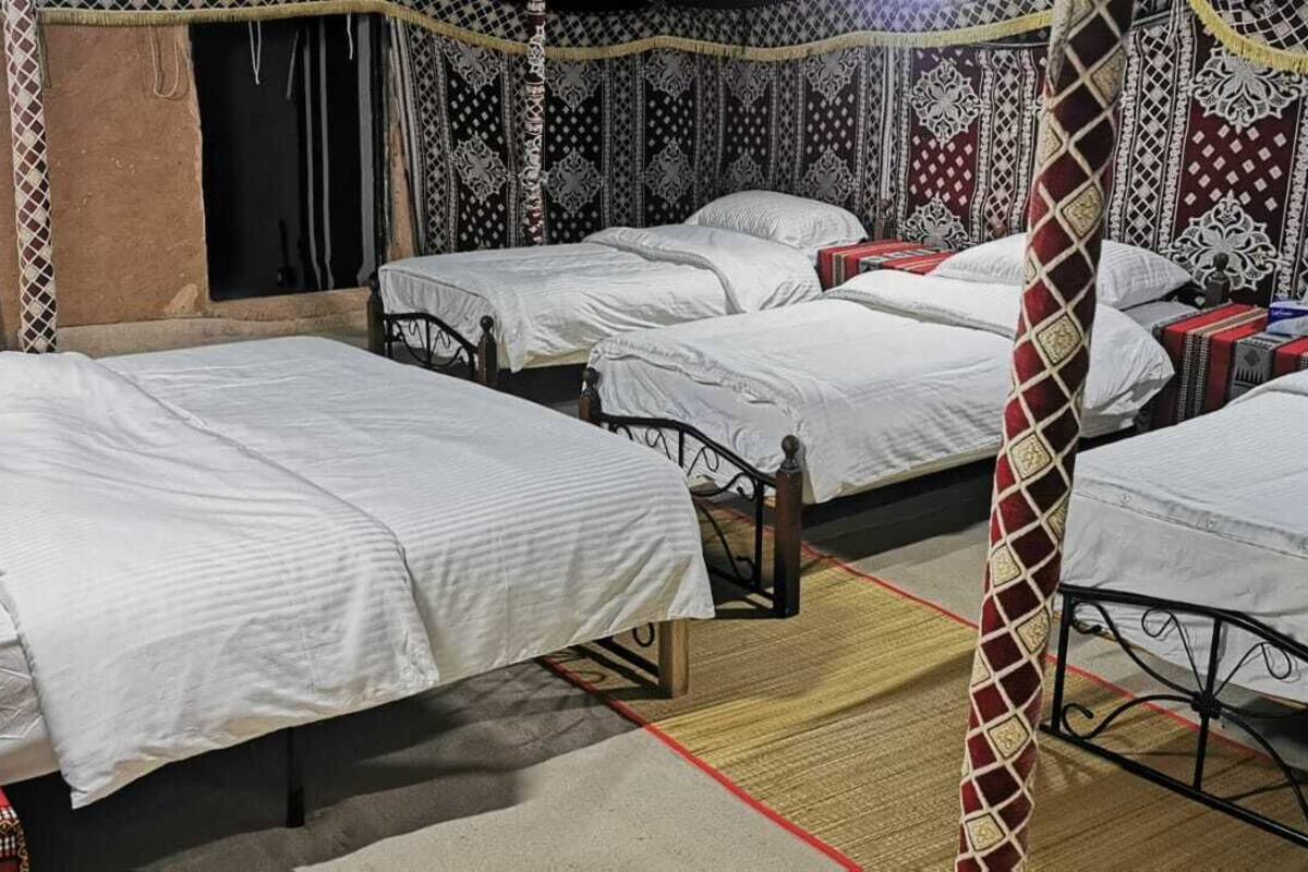 A room with four beds arranged in a row, each with white bedding. There is a patterned rug on the floor, and the walls are decorated with intricate designs.
