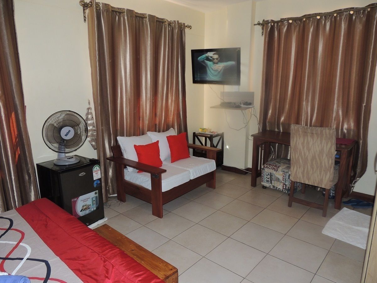 A hotel room with a bed, a sofa, a chair, a TV, and a fan.