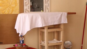 Ein Badezimmer mit einem Holzkonsolenunterschrank, einem weißen Waschbecken, einem Spiegel, einem roten Hocker, einem kleinen runden Tisch mit einer Flasche und einem an der Wand lehnenden Besen.