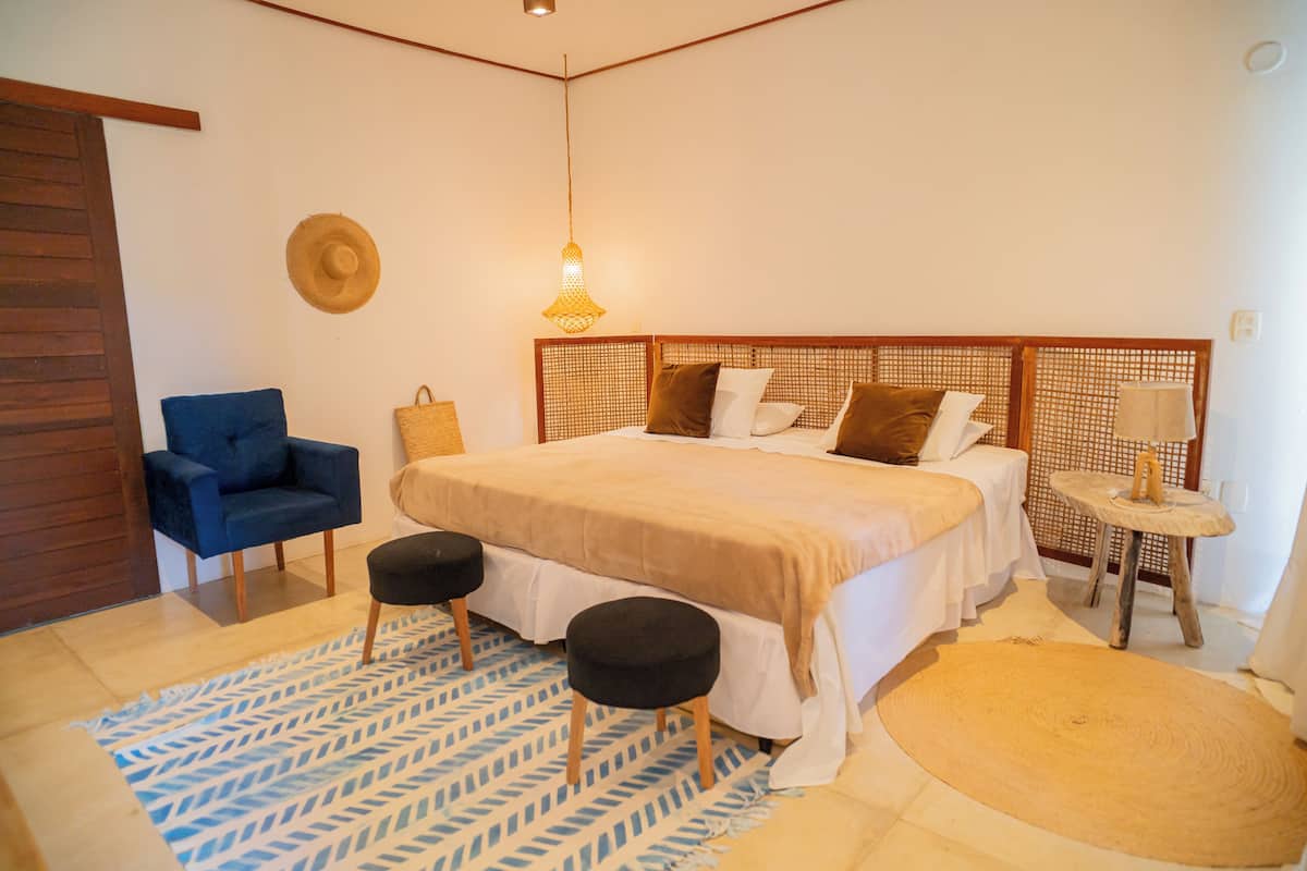 Ein Schlafzimmer mit einem Bett, einem blauen Stuhl, einem kleinen Hocker, einem gewobenen Kopfteil und einem Nachttisch mit einer Lampe.
