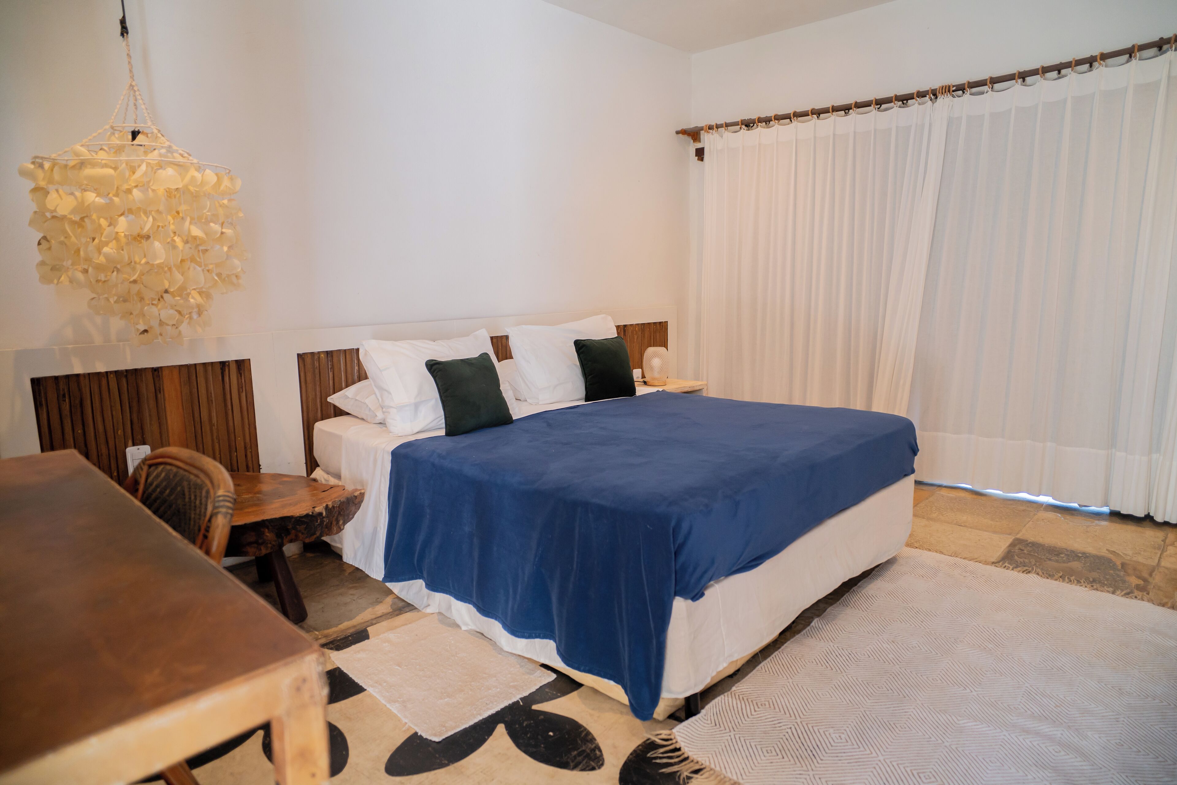 A bedroom with a bed, a wooden table, a hanging light fixture, and a large window with curtains.