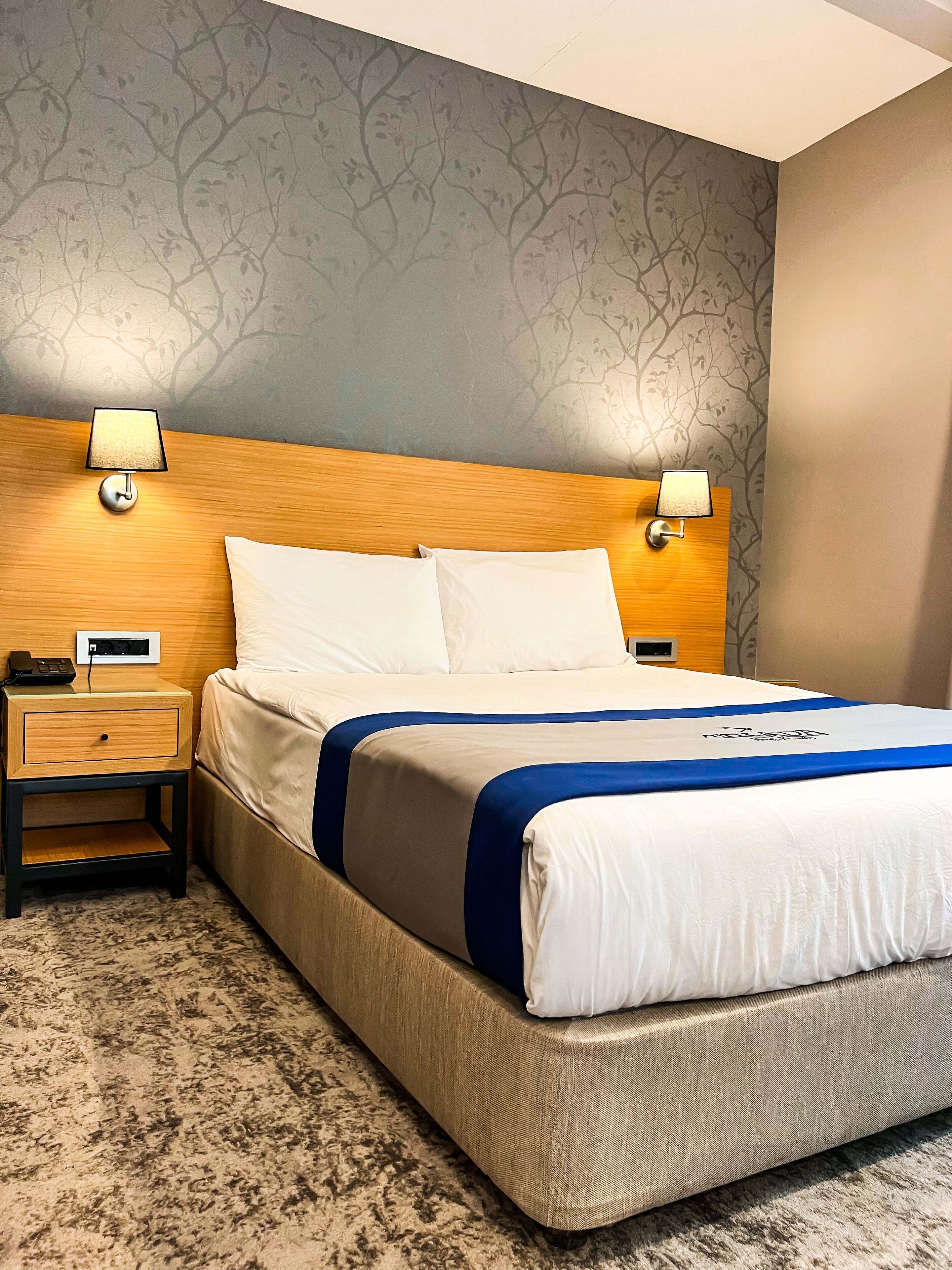 Habitación de hotel con una cama, dos mesitas de noche, una lámpara y un papel tapiz estampado.