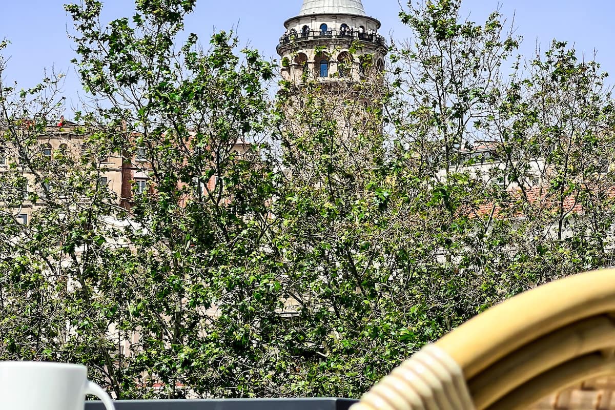 A view of a tower with a conical roof, surrounded by trees, and a wicker chair with a white cup and saucer in the foreground.