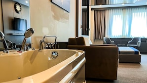 Une salle de bain moderne avec une grande baignoire, un lavabo doté d’un pommeau de douche à main et un miroir. Le salon adjacent comprend un canapé et un téléviseur.