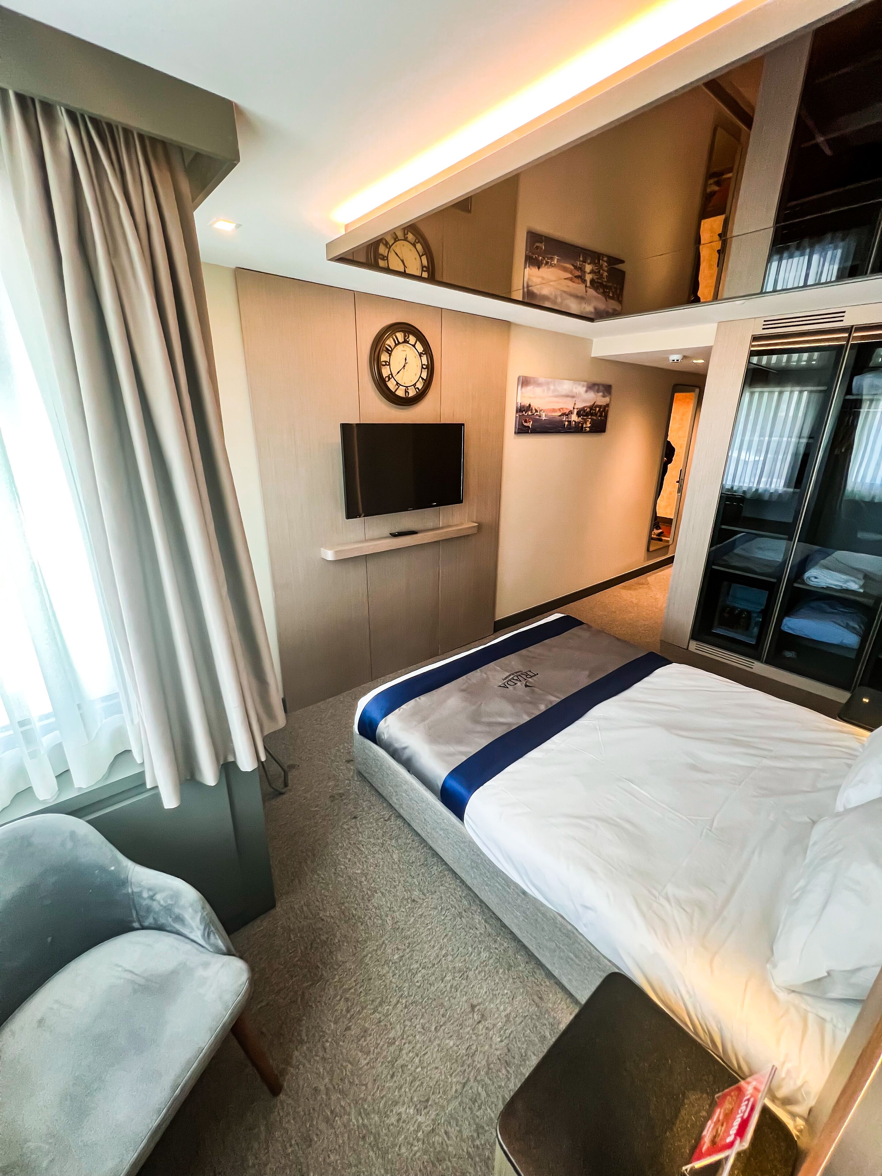 A hotel room with a bed, a TV, a clock, a wardrobe, and a seating area.