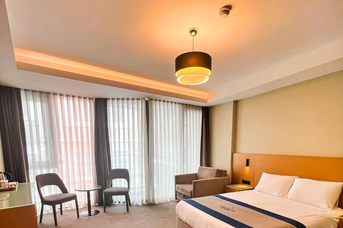 A hotel room with a large bed, a desk, two chairs, a small table, and a large window with curtains.