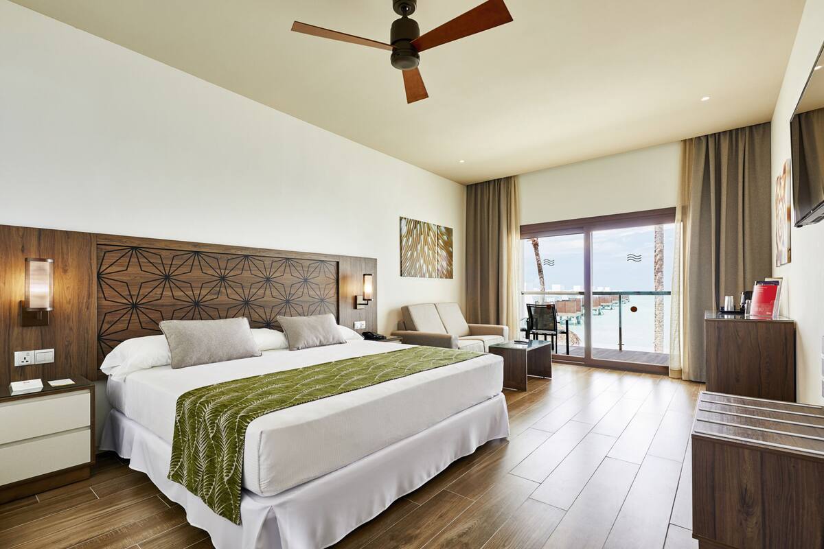 A hotel room with a large bed, a ceiling fan, a desk, a chair, and a view of the city through large windows.