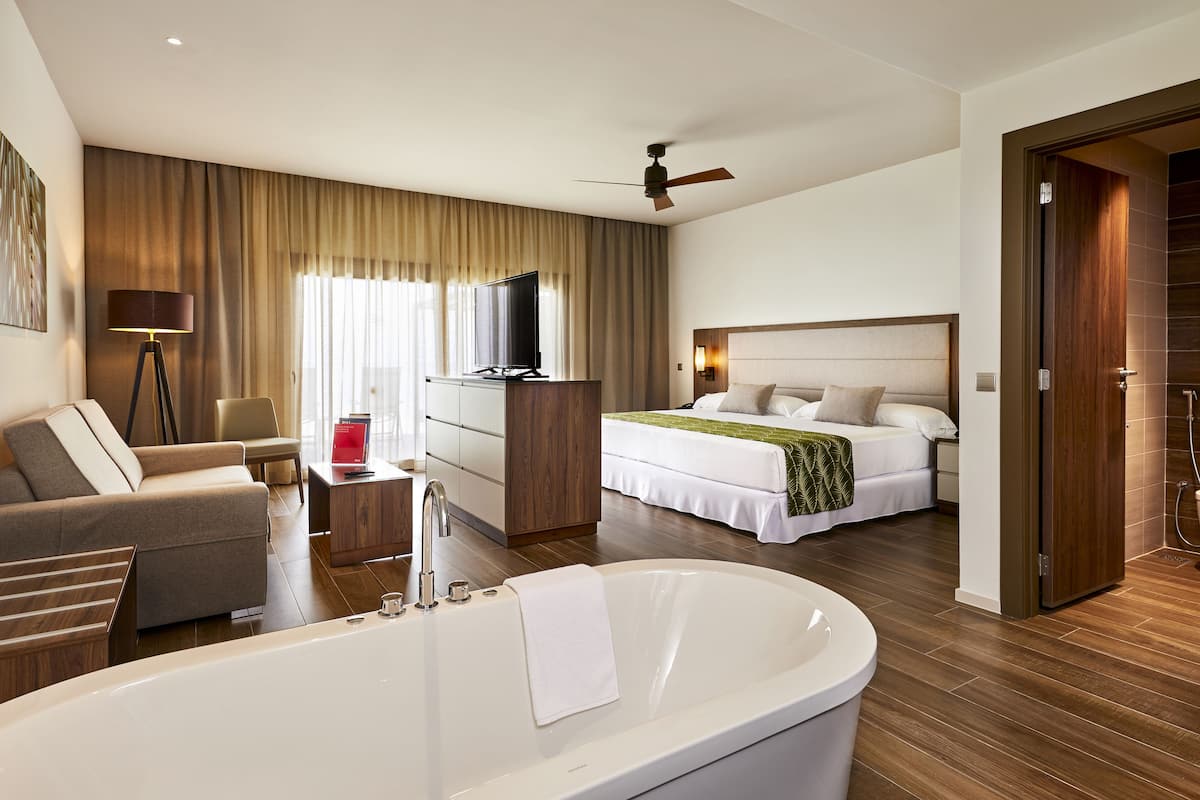 A modern hotel room with a large bathtub, a sofa, a desk with a computer, a television, and a bed with a headboard.