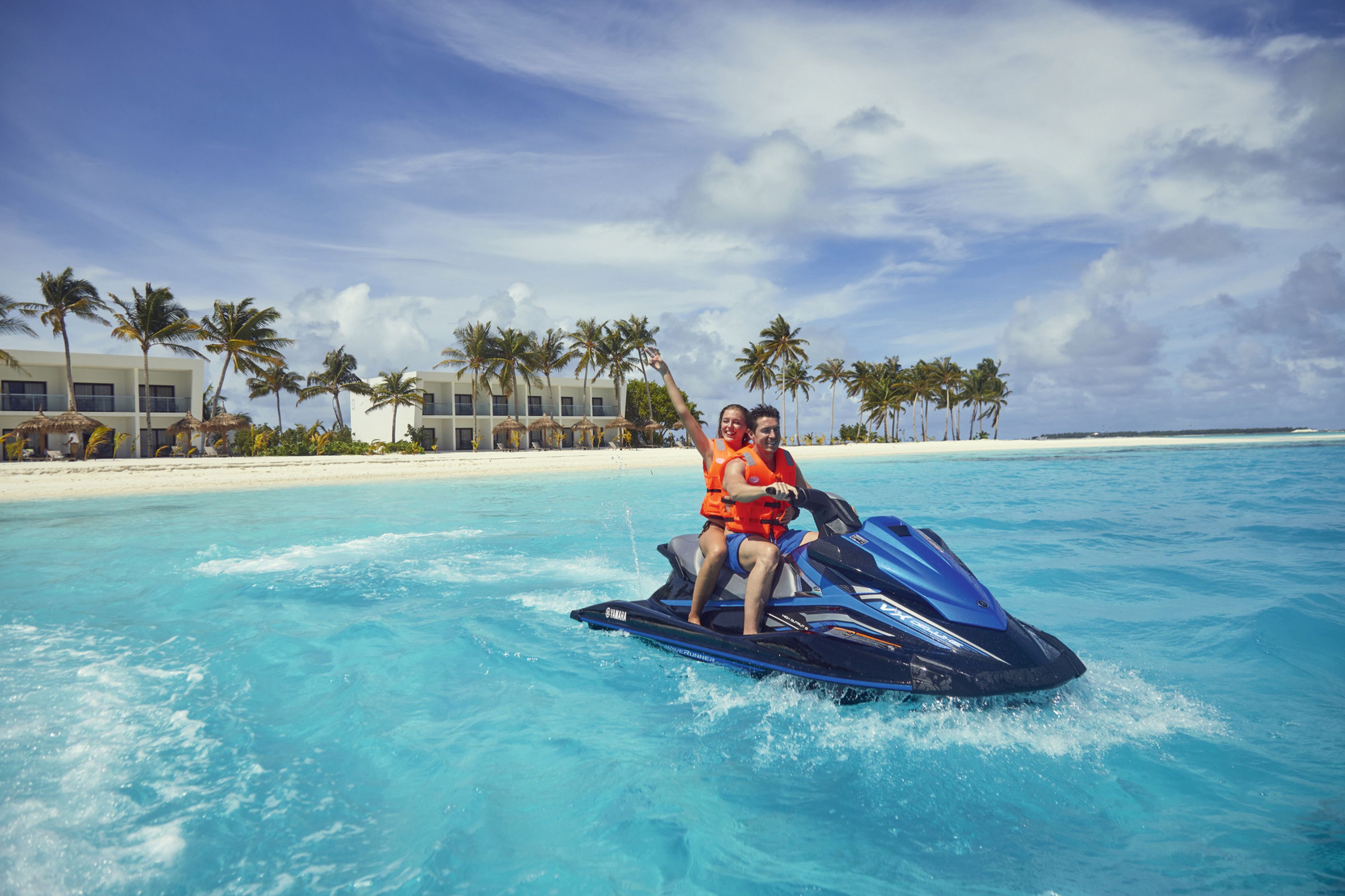 A couple on a jet ski in clear turquoise waters with a resort in the background.