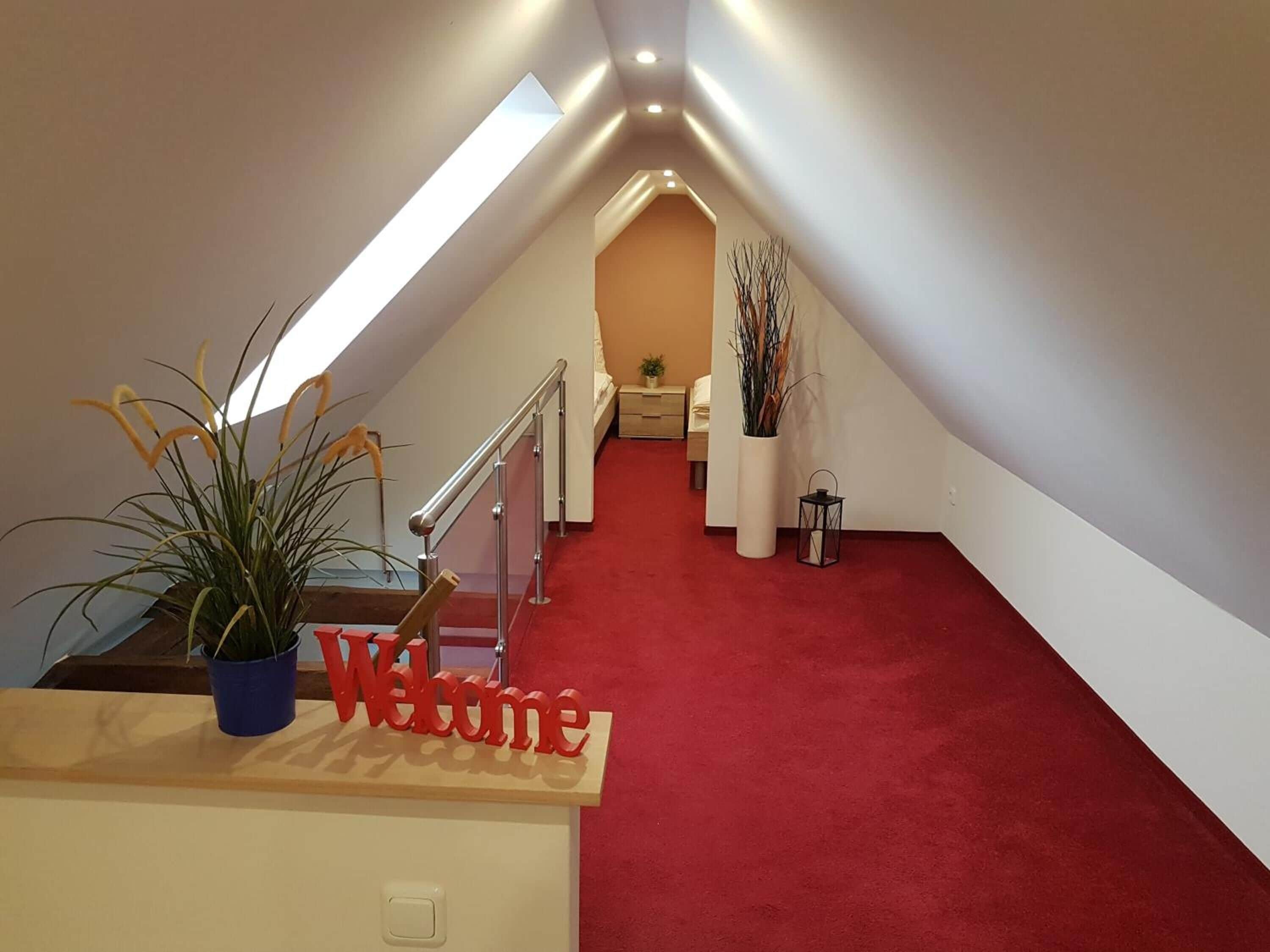 A red carpeted hallway leading to a bedroom with a bed, a nightstand, and a potted plant. A 'Welcome' sign is visible on the left.