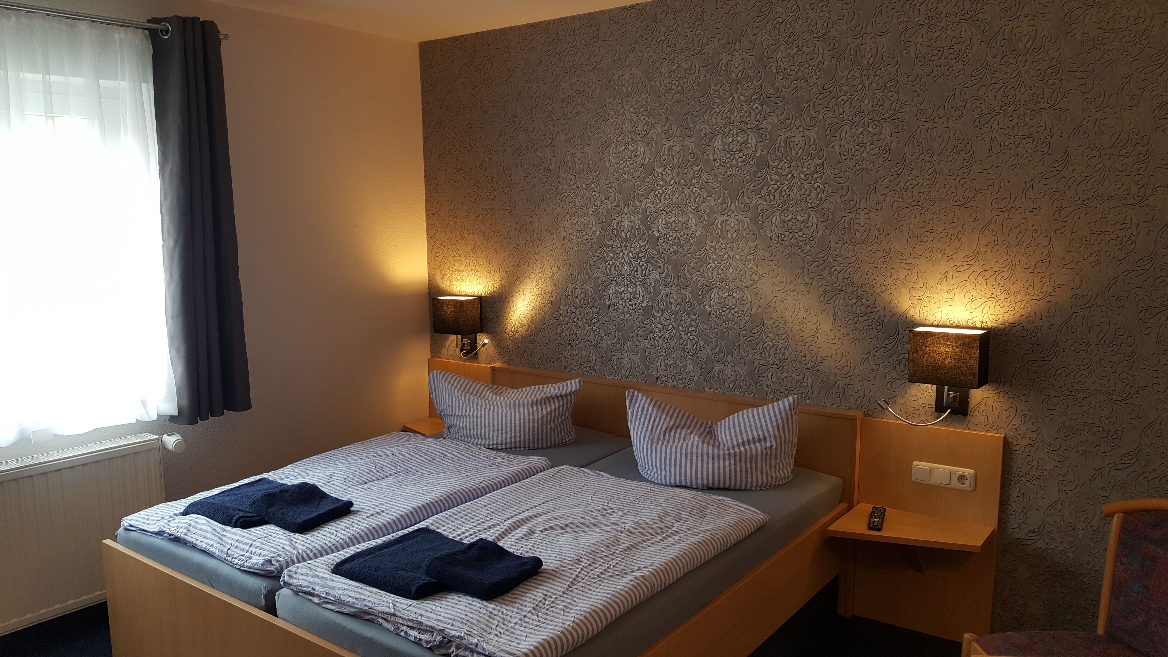 A hotel room with a bed, two bedside lamps, a window with curtains, and a patterned wall.