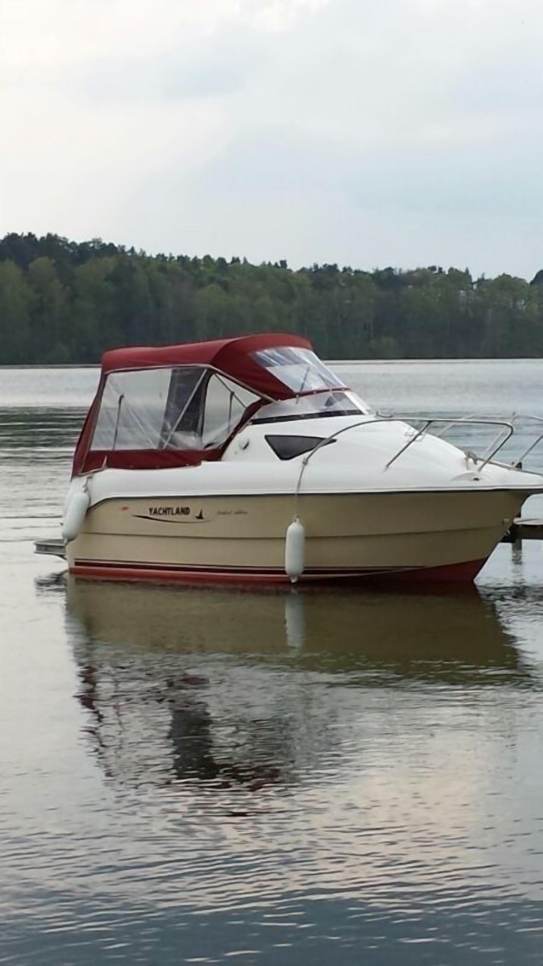 A white and red boat named "YACHTLAND" on calm water with a forested shoreline in the background.