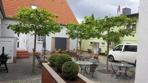 Een terras met tafels en stoelen, omringd door groen en een wit gebouw met een rood dak.