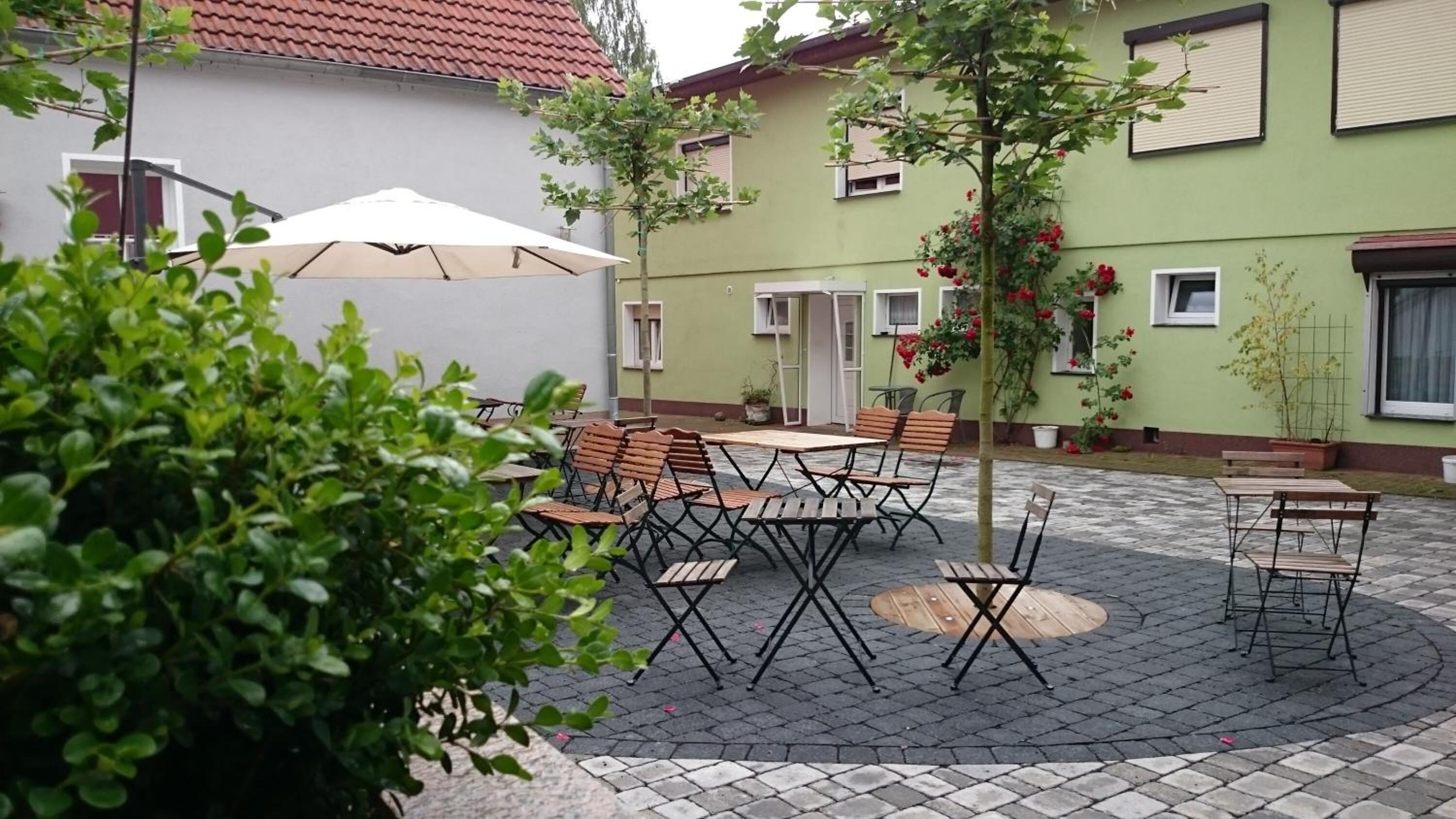 Une terrasse extérieure avec des tables et des chaises, entourée de verdure et bordée d’un bâtiment dont le toit est recouvert de tuiles.