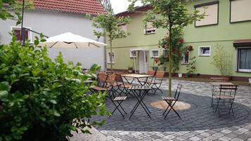 Une terrasse extérieure avec des tables et des chaises, entourée de verdure et bordée d’un bâtiment dont le toit est recouvert de tuiles.