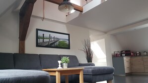 Een moderne woonkamer met een donkergrijze bank, een houten salontafel en een potplant. Aan de muur hangt een groot gepoft schilderij van een pier, en er is een keuken te zien op de achtergrond.