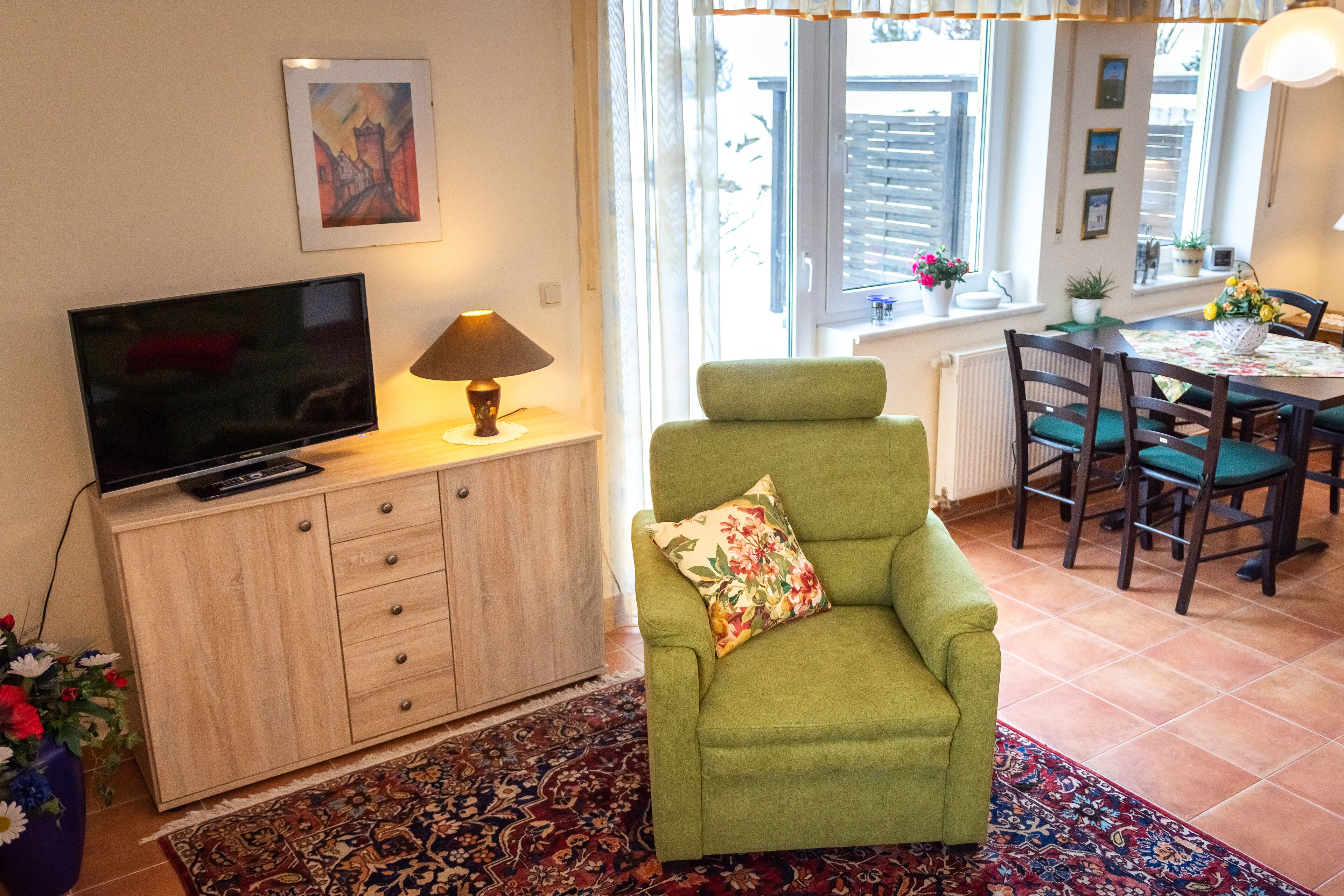 A living room with a green armchair, a flat-screen TV on a wooden cabinet, a patterned rug, and a dining area with a table and chairs.