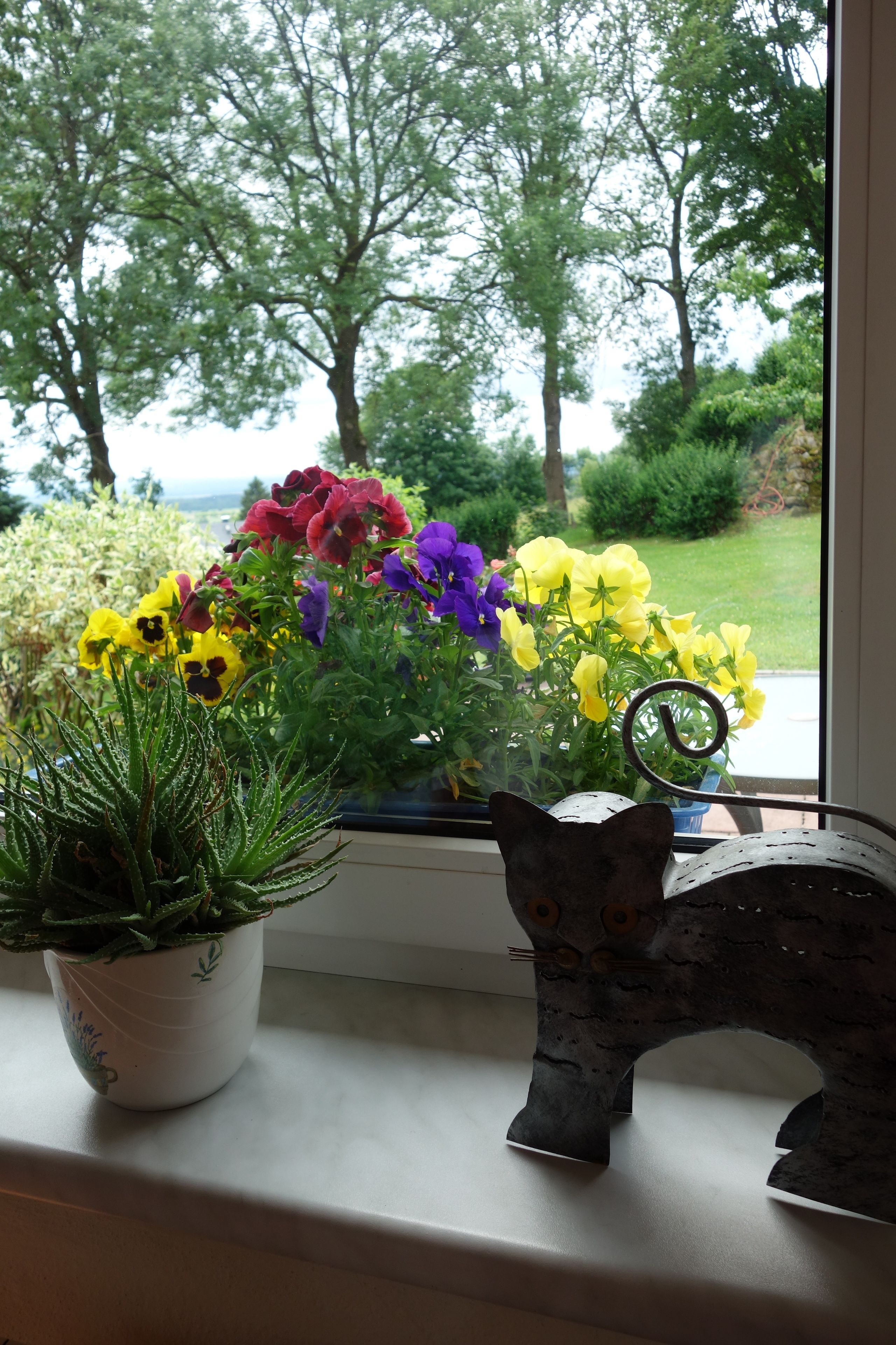 A window sill with a potted plant, a metal sculpture, and a window with a view of trees and a grassy area.