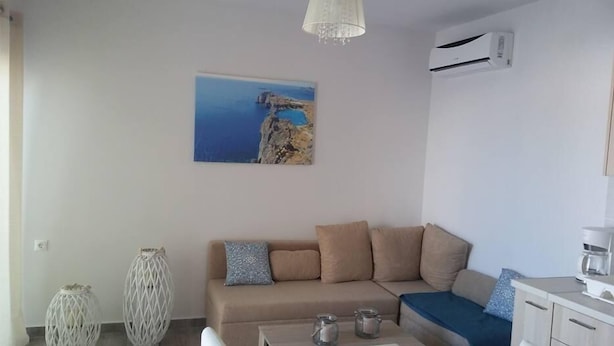 A living room with a beige sofa, a coffee table, a painting of a coastal landscape, and a white air conditioning unit. - Nianthy Apartments (Rhodes)