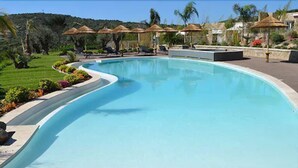 Una piscina grande y curvada con sillas de descanso y sombrillas, rodeada de jardines ajardinados y colinas.
