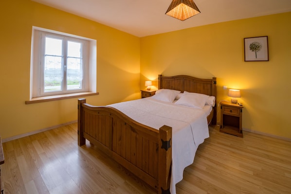 Un dormitorio con una cama de madera, dos mesitas de noche con lámparas, una ventana con vistas y un cuadro enmarcado en la pared.