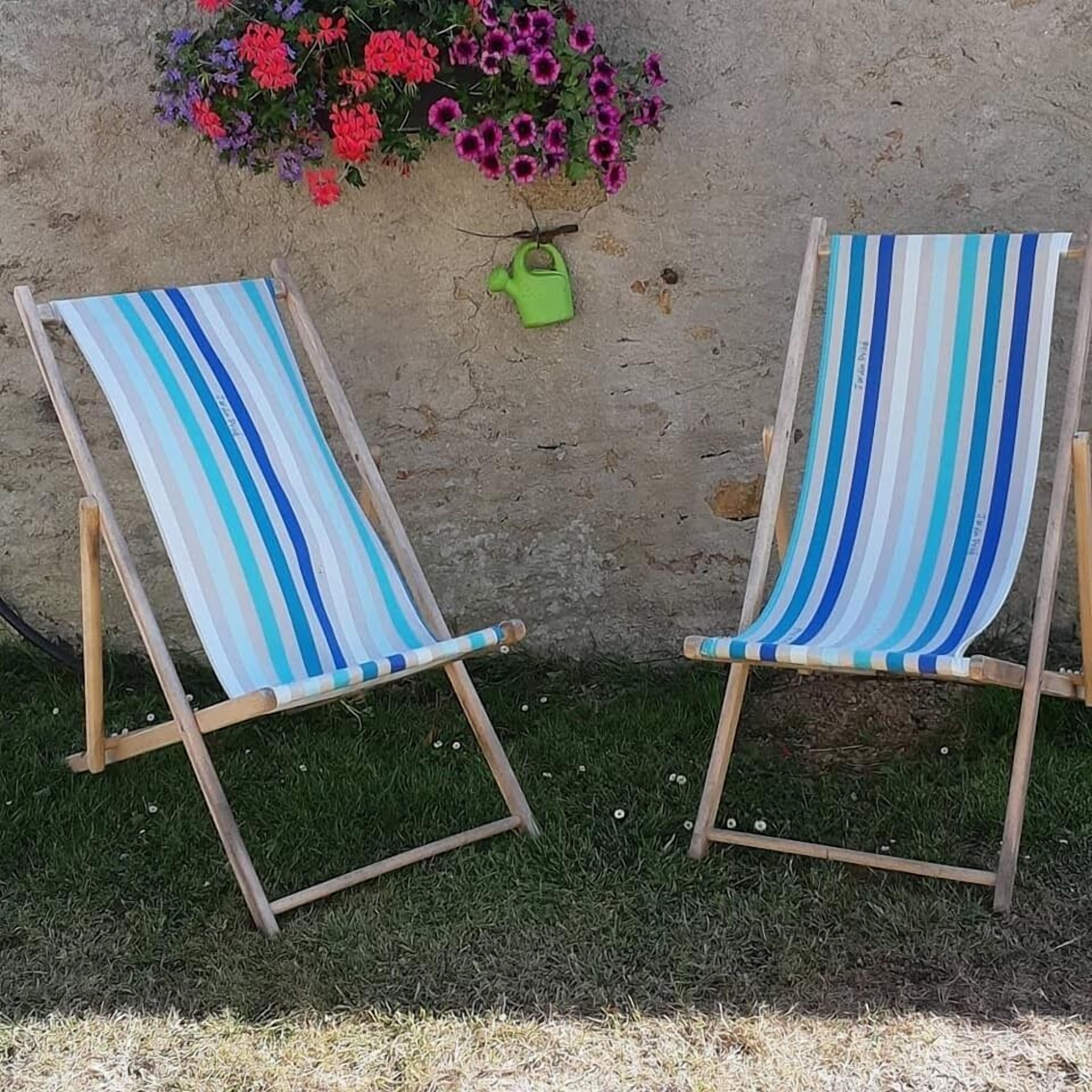 Deux chaises de plage à rayures, avec une structure en bois et un siège en tissu, sont placées sur l’herbe, adossées à un mur sur lequel est posé un pot de fleurs.