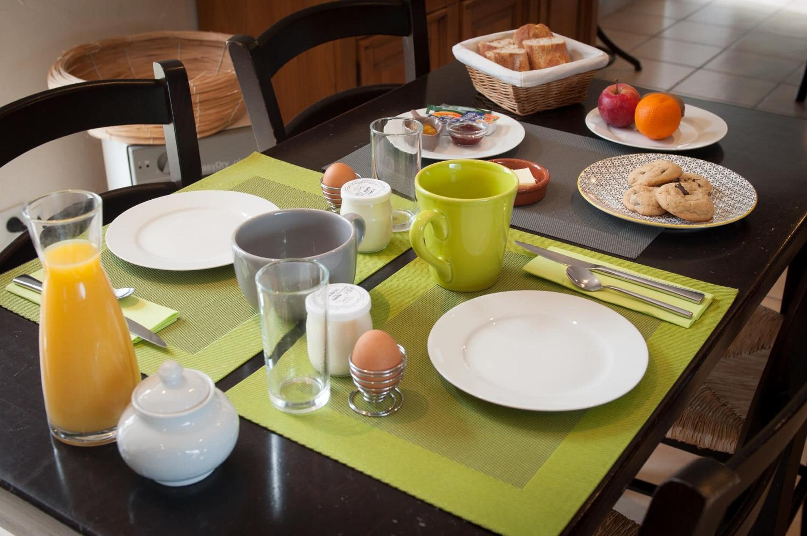 Una mesa de desayuno con platos, una taza, un vaso de jugo de naranja, una cesta de pan y un plato con galletas.