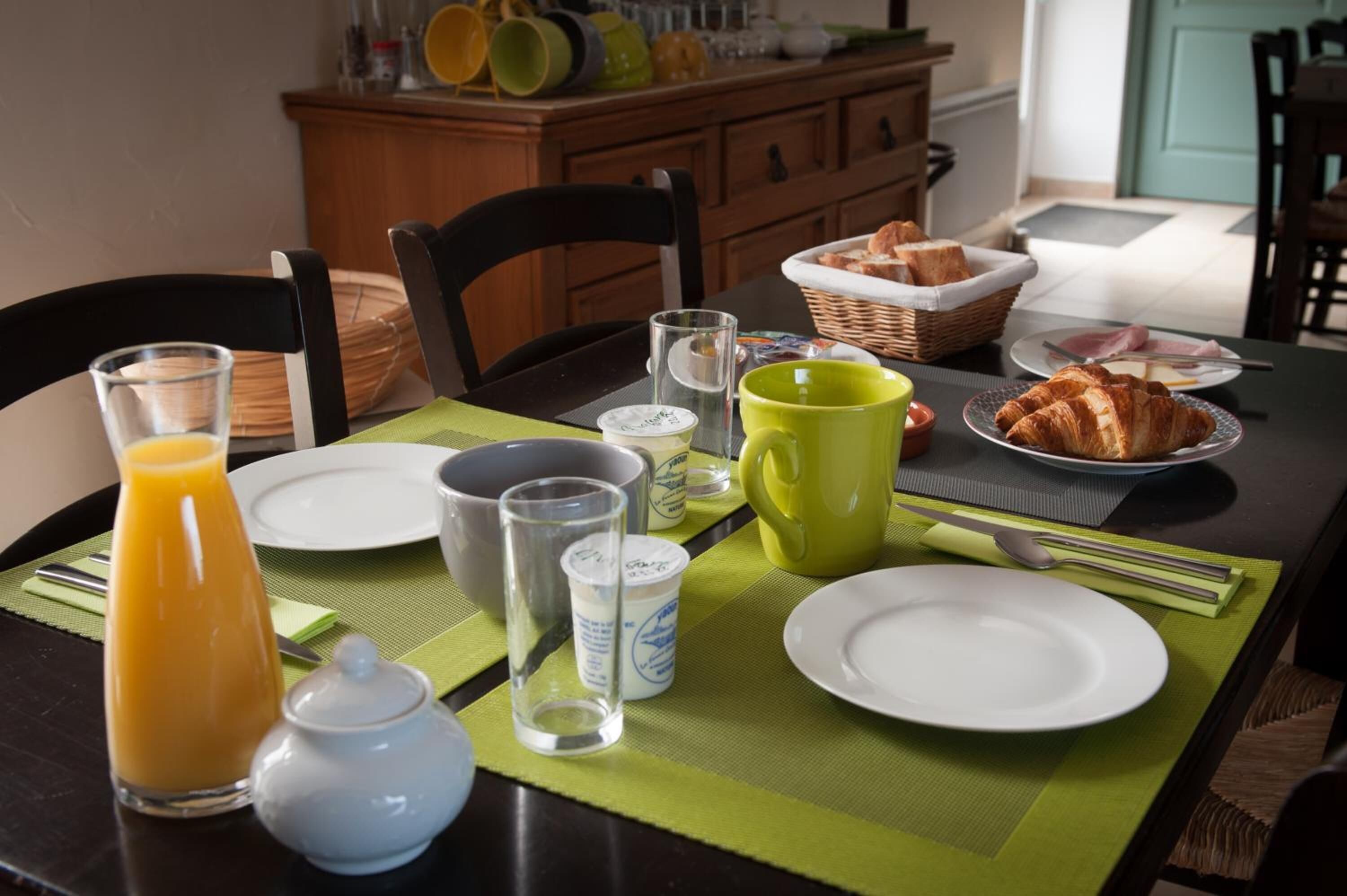 Een ontbijttafel gedekt met borden, een glas sinaasappelsap, een gele mok en een mandje croissants.