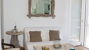 Une pièce avec un canapé blanc, deux coussins, une table basse ronde en bois sur laquelle se trouvent des bols et un bocal en verre, une petite table ronde ornée d’objets décoratifs et un miroir avec un cadre rustique.