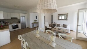 Une cuisine spacieuse équipée d’appareils électroménagers modernes, un coin repas avec une table et des chaises en bois, et un salon confortable avec un canapé et une grande fenêtre.